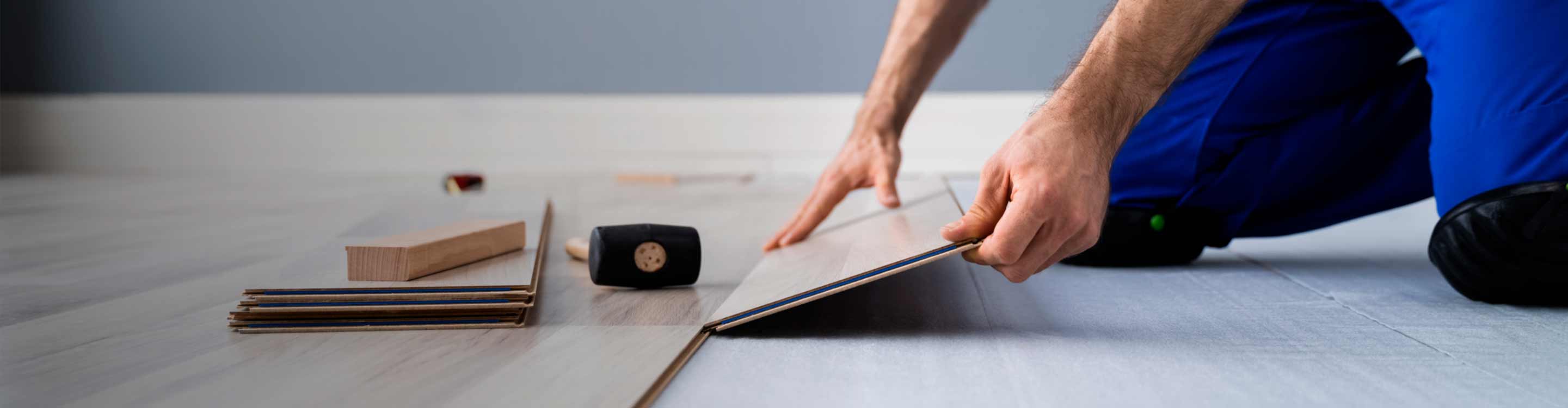 Close-up of a person installing light wood-grain flooring, using a rubber mallet and tapping block, with tools and planks visible in a well-lit space.